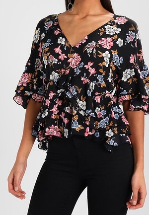 Blouse cache-cœur à col en V noire avec imprimé floral rose, blanc et bleu, manches volantées et ourlet à basque portée avec un pantalon noir.