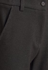 Gros plan sur un pantalon gris foncé texturé avec de fines lignes diagonales, une ceinture boutonnée et une poche latérale visible.