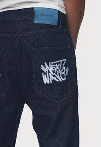 Mörkblå denimjeans med en texturerad yta, som har en broderad "NEXT WAY"-logotyp på bakfickan och en blå tygetikett med texten "PEGADOR."