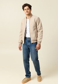 Beige bomberipuku, valkoinen T-paita, siniset farkut, beige mokkasiniset kengät. Takissa on pyöreä kaula-aukko, ribbineulekädensuut ja sileä pinta.