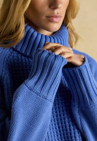 Niebieski sweter z golfem wykonany z dzianiny, charakteryzujący się teksturowanym wzorem, ściągaczami u dołu rękawów oraz luźnym krojem. Materiał wygląda na miękki i ciepły.