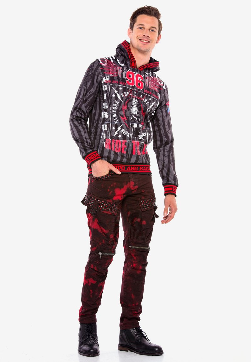 Schwarzer und roter Hoodie mit Grafikdruck, hohem Kragen und gerippten Bündchen. Dunkelrote Hose mit Spritzer-Muster und Nietenakzenten. Schwarze Stiefel.