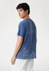 Camiseta de manga corta azul con un patrón de paisley en la mitad inferior, con "GUESS" impreso en un tono más claro en la parte posterior. Material de algodón.