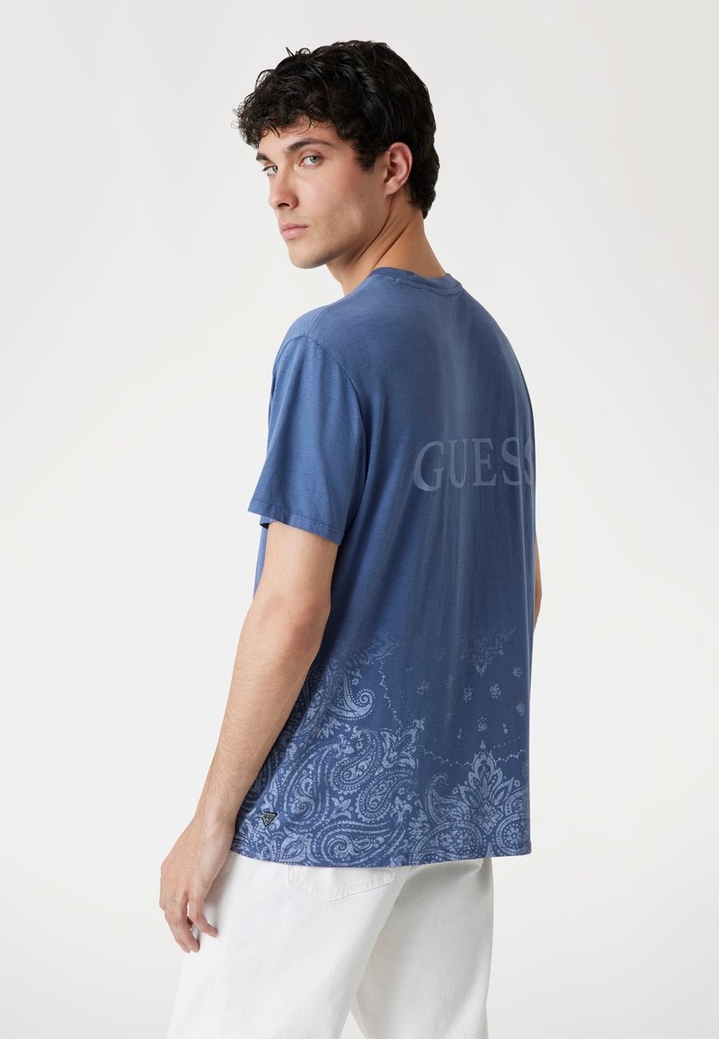 Camiseta de manga corta azul con un patrón de paisley en la mitad inferior, con "GUESS" impreso en un tono más claro en la parte posterior. Material de algodón.