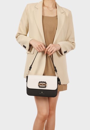 Donna con blazer beige e vestito marrone che tiene in mano una borsa nera e bianca con fibbia intrecciata e chiusura dorata davanti a uno sfondo chiaro.