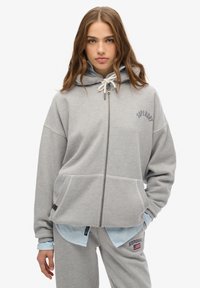Graue Zip-Hoodie aus weicher Baumwolle, mit geräumiger Passform, zwei Fronttaschen und einem gestickten Logo auf der Brust. Kapuze mit Kordelzug.