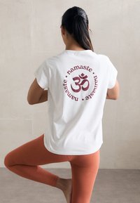 Bijela pamučna majica s crvenim kružnim uzorkom koji sadrži riječ "namaste" i simbol "Om" na leđima. Okrugli izrez, kratki rukavi, opušteni kroj.