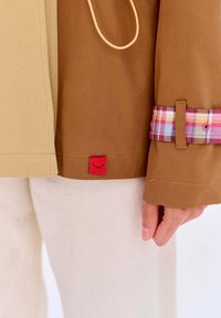 Hnedo-bežová bunda s károvaným manžetovým detailom a červenou nášivkou s grafikou zatvoreného oka. Hladká látka, oranžový šnúrka na stiahnutie a dostatok švy.