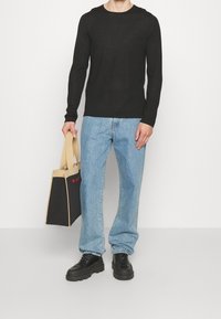 Person in einem schwarzen Langarmshirt, hellblauen Jeans, schwarzen klobigen Schuhen, der eine schwarze und beigen Tragetasche mit roter Schrift hält.