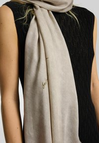 Beige scarf draperet omkring halsen, lavet af blødt stof med en subtil glans, med et håndskrevet accent i guld. Båret sammen med en struktureret sort top.