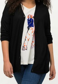 Schwarze Strickjacke mit langen Ärmeln und Knopfschluss. Darunter ein weißes Hemd mit buntem grafischen Design, kombiniert mit blauen Jeans.