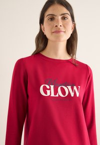 Červená košile s dlouhými rukávy vyrobená z měkké tkaniny, na které je bílé písmo "Mountain GLOW" a "REACH HEIGHTS" v tučném designu.