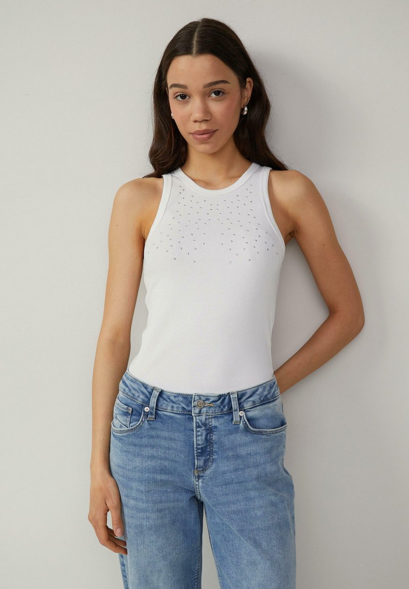 Witte geribbelde mouwloze top met strasstenen accenten aan de voorkant; gecombineerd met lichtblauwe high-waisted denim jeans.