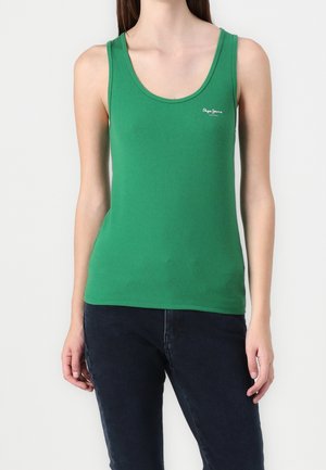 Kobieta w zielonym, prążkowanym, bezrękawniku z małym białym logo na piersi, w połączeniu z ciemnoniebieskimi dżinsami.