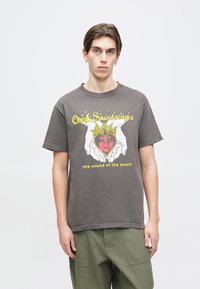 Camiseta de algodón gris con mangas cortas. Gráfico colorido de manos sosteniendo un corazón con llamas, con el texto "Obey Soundscapes" debajo.