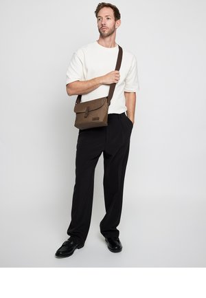 Hombre con camisa blanca de manga corta, pantalones negros, zapatos negros, sujetando la correa de un bolso marrón cruzado, de pie frente a un fondo claro y sencillo.