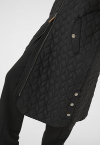Veste matelassée noire avec une fermeture éclair latérale, présentant un motif en losanges, des boutons-pression le long de l'ourlet, et une finition en tissu lisse.