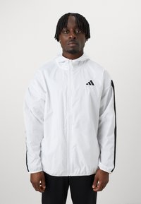 Casaco corta-vento branco com riscas laterais pretas, fecho à frente e capuz. Apresenta três riscas diagonais pretas com o logo no lado superior esquerdo do peito.