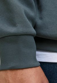 Gray sweatshirt sleeve with ribbed cuffs, showcasing smooth fabric texture and a neat finish. Underneath, a white shirt hem is visible.