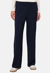 Pantalons larges bleu marine, fabriqués dans un tissu lisse et extensible avec une taille plate élastique. Associés à des baskets blanches.