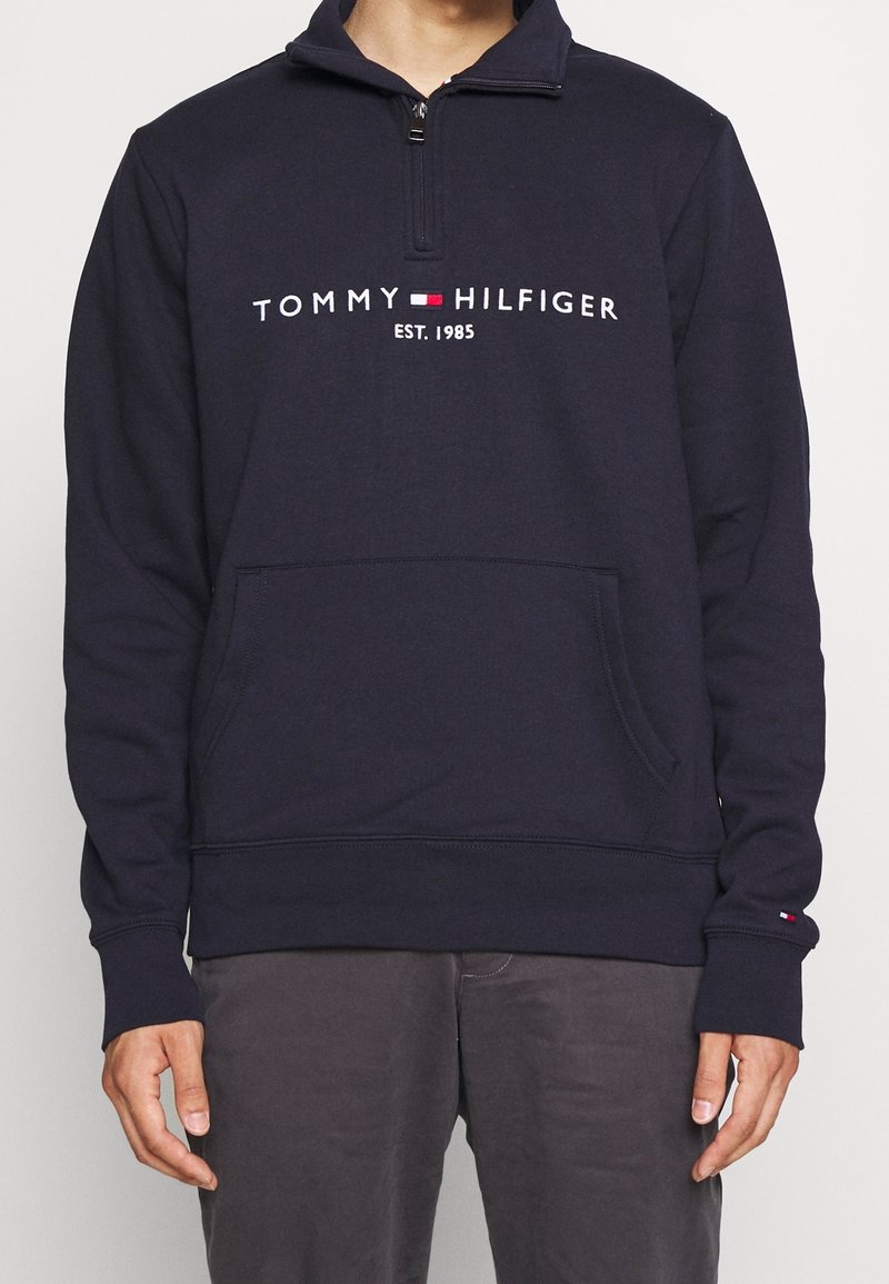 Sweat à capuche Tommy Hilfiger bleu marine avec poche avant et col à fermeture éclair, avec logo de la marque et texte "EST. 1985" sur la poitrine.