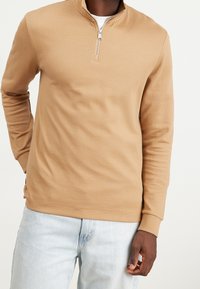 Pullover in cotone color tan con zip a un quarto, realizzato in tessuto testurizzato, dotato di maniche lunghe e dettagli della zip in contrasto. Abbinato a jeans azzurri.