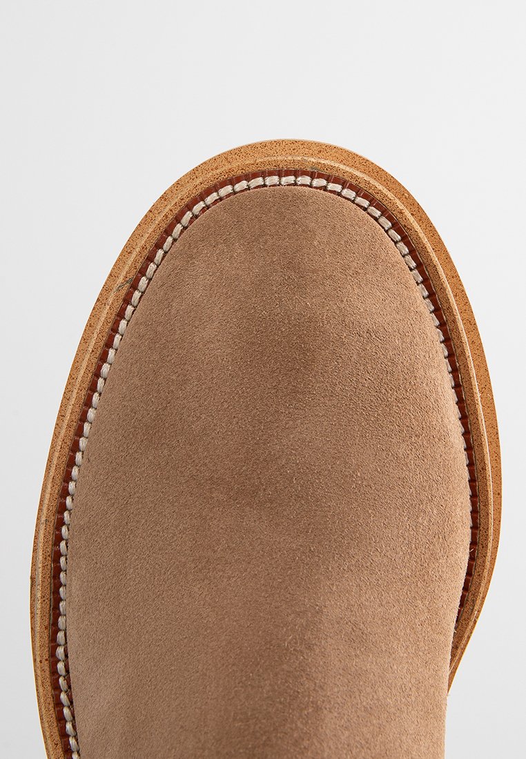 Braune Wildlederstiefel mit einer runden Spitze, ausgestattet mit einer genähten tanfarbenen Gummisohle und kontrastierenden weißen Nähte entlang des Randes.