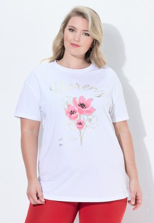 Blondynka w białym T-shircie z różowym kwiatowym nadrukiem i złotym napisem "Florenz", w połączeniu z czerwonymi spodniami, stoi na białym tle.