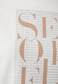 Witte T-shirt met een groot bruine "S" en meerdere kleine grijze teksten "SELFCONFIDENT" in een herhalend patroon. Zacht, katoenmateriaal.