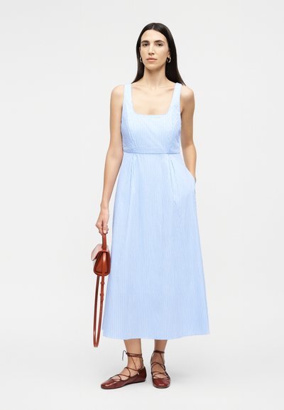 Femme aux longs cheveux bruns portant une robe midi sans manches à rayures bleu clair, des chaussures plates marron à lacets, et tenant un sac à main marron.