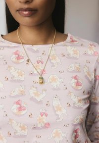 Een close-up van een vrouw die een lichtpaarse shirt draagt met een speels kattenpatroon en een gouden ketting met een klein hangertje.