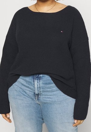 Persona vistiendo un suéter de punto negro con un pequeño logo en el pecho y jeans de cintura alta color azul claro, posando sobre un fondo liso.