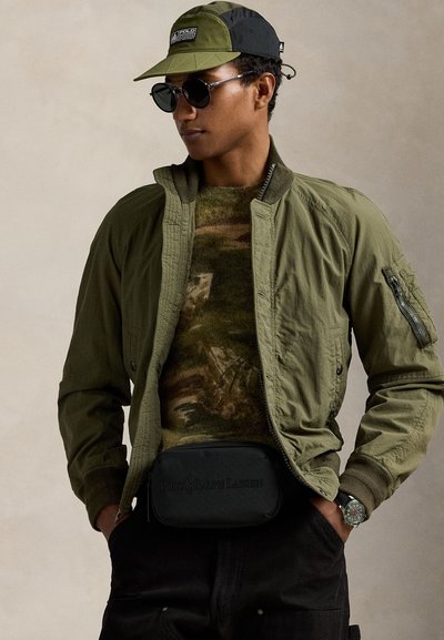 Vīrietis valkā olīvu zaļu bombera jaku, kamuflāžas džemperi, zaļas un melnas krāsas cepuri, melnas saulesbrilles, melnas bikses, pulksteni un melnu Polo Ralph Lauren vidukļa somiņu.