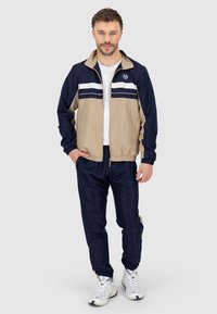 Tuta leggero con zip in beige e blu navy, caratterizzato da strisce bianche sul petto, con polsini elasticizzati e vita regolabile.