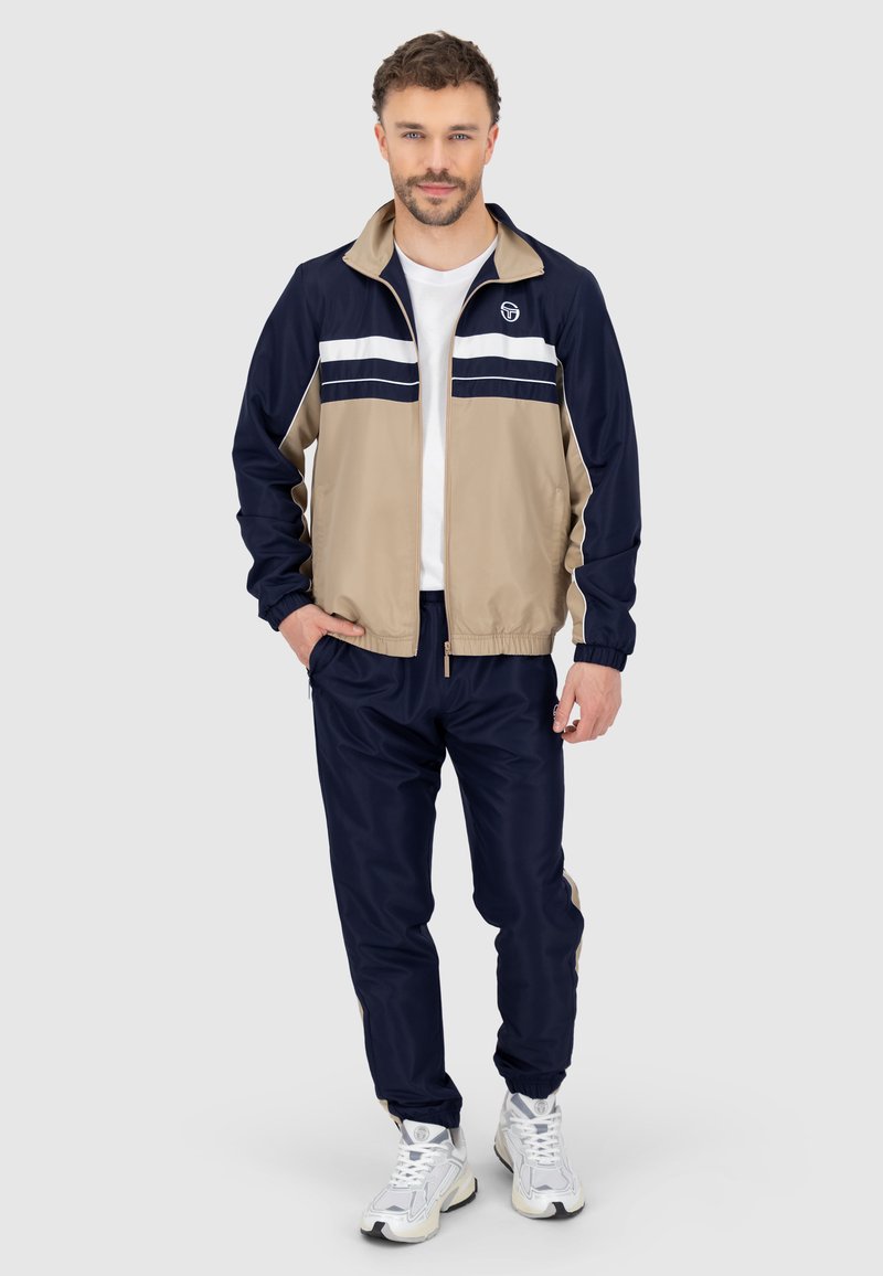 Tuta leggero con zip in beige e blu navy, caratterizzato da strisce bianche sul petto, con polsini elasticizzati e vita regolabile.