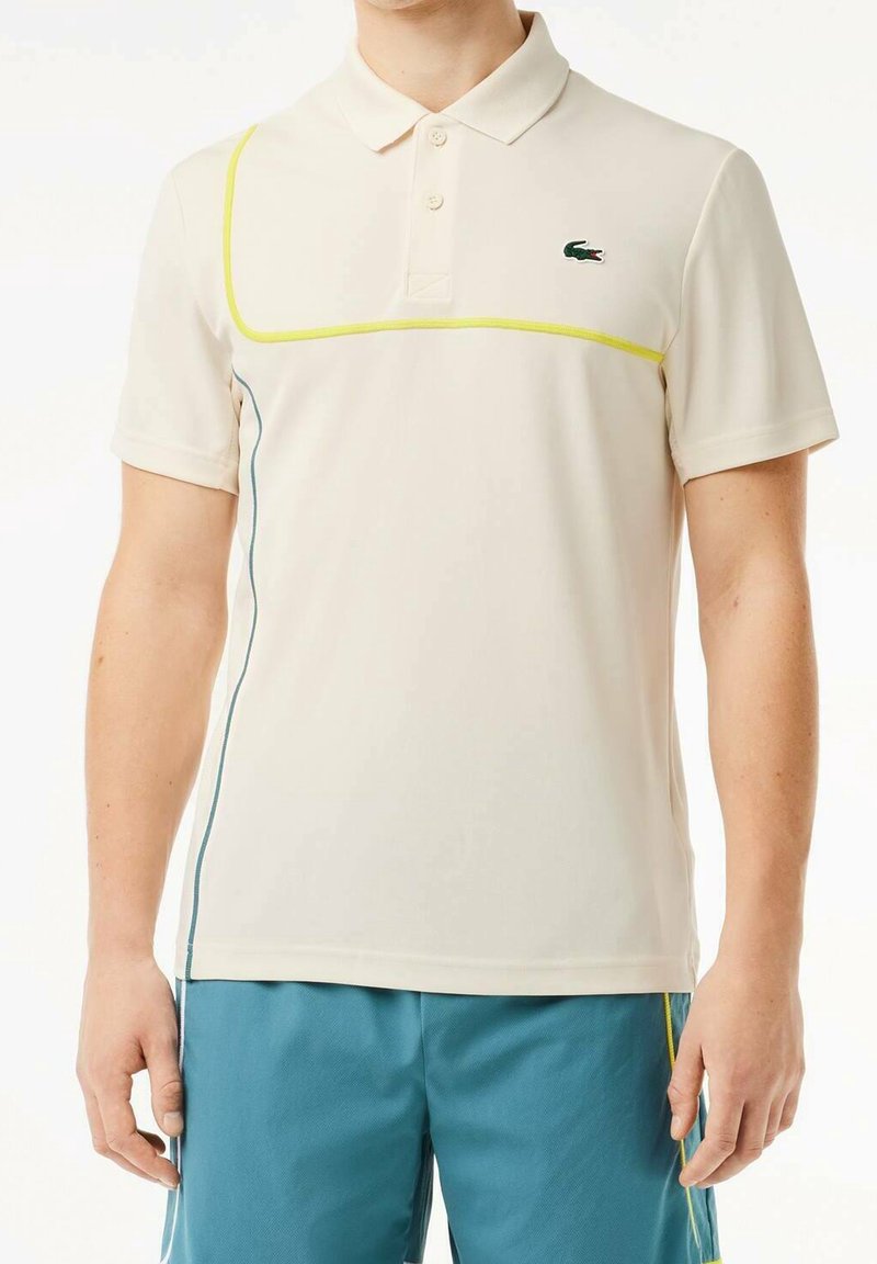 Homme portant un polo crème avec des passepoils jaunes et bleus et un petit logo crocodile vert, associé à un short bleu.