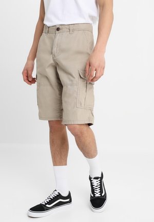 Mann trägt beige Cargo-Shorts, weiße Sportsocken, schwarze und weiße Sneaker sowie ein weißes T-Shirt und steht vor einem einfarbigen Hintergrund.