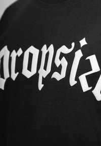 T-shirt noir en coton avec une inscription en lettres gothiques blanches audacieuses qui épellent "dropsie". La vue rapprochée met en évidence la texture et le design.