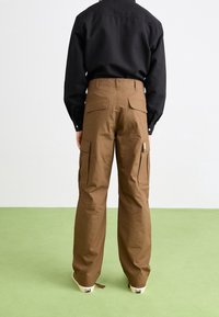 Braune Cargo-Hosen mit einem lockeren Schnitt, ausgestattet mit mehreren Taschen und einer glatten Textur. Getragen mit einem eng anliegenden schwarzen Shirt und lässigen Schuhen.