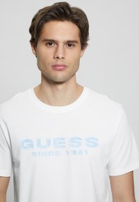 Witte katoenen T-shirt met een lichtblauw, glinsterend "GUESS" logo en de tekst "SINCE 1981", ronde halslijn en korte mouwen.