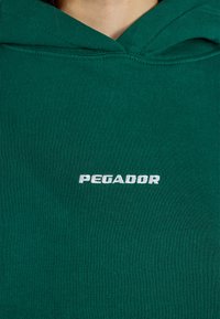 Hoodie verde em algodão com um bolso frontal e capuz ajustável por cordão. Apresenta "PEGADOR" bordado em branco na parte superior do peito.