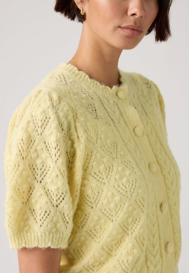 Woman wearing a pale yellow knitted buttoned cardigan with a diamond lace pattern and short puff sleeves.