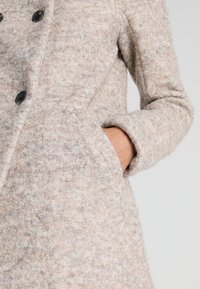 Cappotto in lana beige chiaro con finitura testurizzata, dotato di grandi bottoni neri e tasche laterali. Maniche lunghe e design sartoriale.
