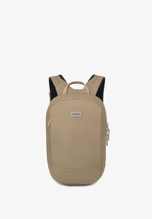 Sac à dos en tissu beige avec une forme rectangulaire, doté de bretelles rembourrées, d'une poignée supérieure et d'une poche zippée à l'avant. Étiquette de marque visible.