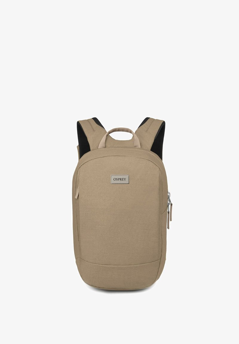 Sac à dos en tissu beige avec une forme rectangulaire, doté de bretelles rembourrées, d'une poignée supérieure et d'une poche zippée à l'avant. Étiquette de marque visible.