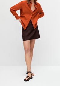 Chemise en lin couleur rouille avec un devant à boutons, associée à une jupe marron foncé. Le modèle porte des sandales noires à lanières. Coupe simple et moderne.