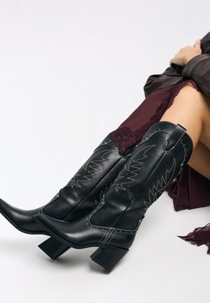 Donna che indossa stivali da cowboy in pelle nera con cuciture decorative, abbinati a un abito bordeaux e a una giacca scura.