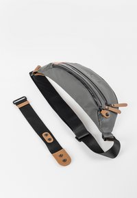 Sac banane en tissu gris avec une forme arrondie, fermeture éclair, accents en cuir beige et une bandoulière noire amovible avec des boutons-pression.