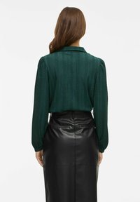 Blusa verde scuro con texture, maniche a sbuffo e colletto, abbinata a una gonna di pelle nera a vita alta.