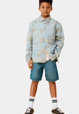 Niño de pie con las manos en los bolsillos, vistiendo una camisa de camuflaje azul grisáceo, pantalones cortos vaqueros, calcetines blancos con rayas negras y zapatillas negras.